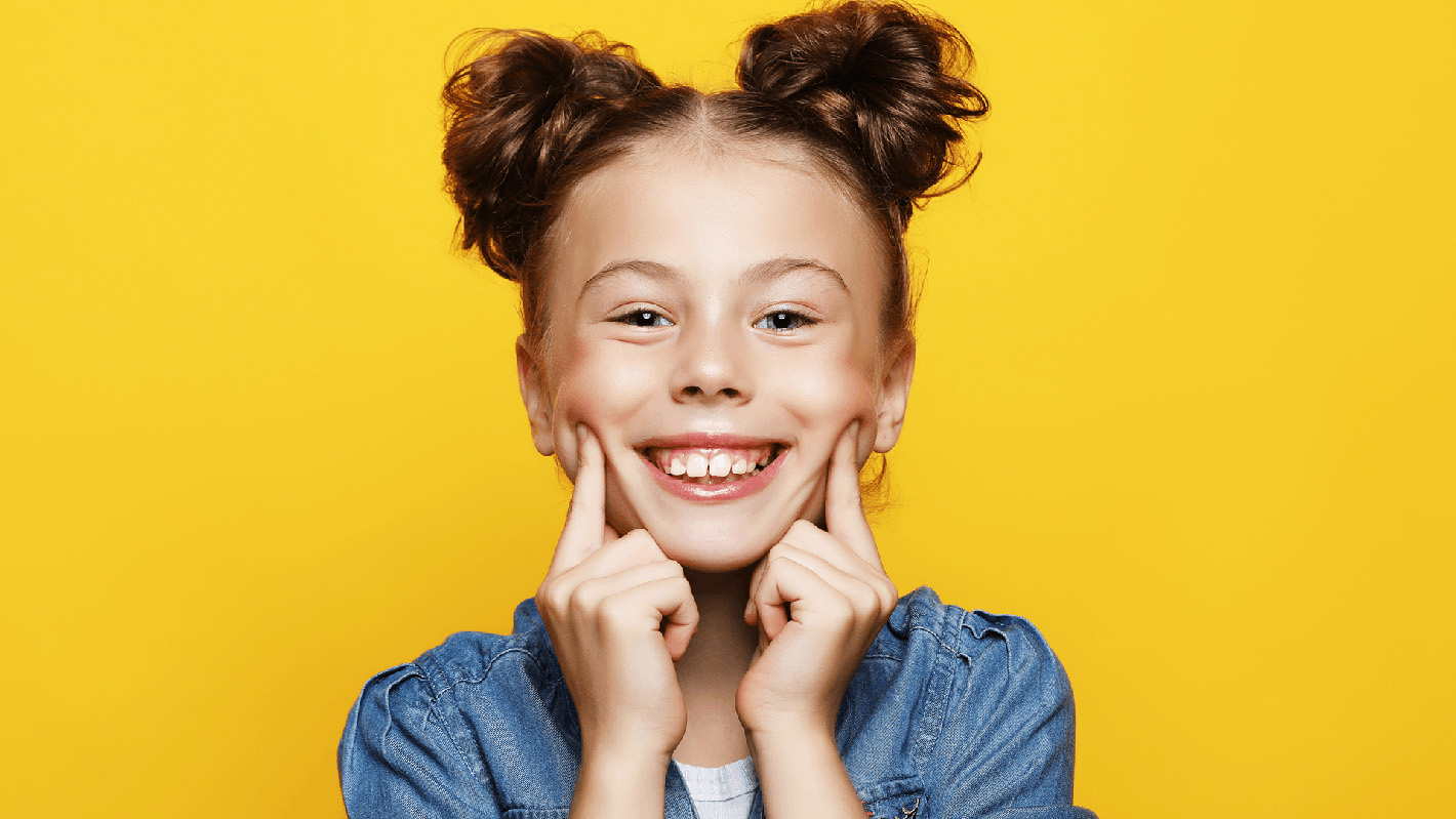 Close up of a young girl who is smiling and holding up her smile with her two pointer fingers. She is wearing a jean jacket and is in front of a yellow background 