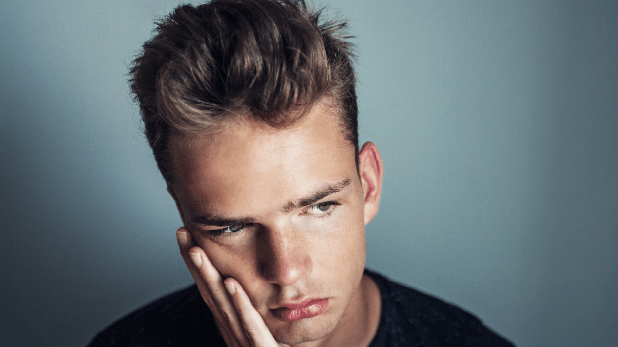 Close up shot of a young blonde mail holding a hand to the right side of his face and looks to be in pain from his teeth