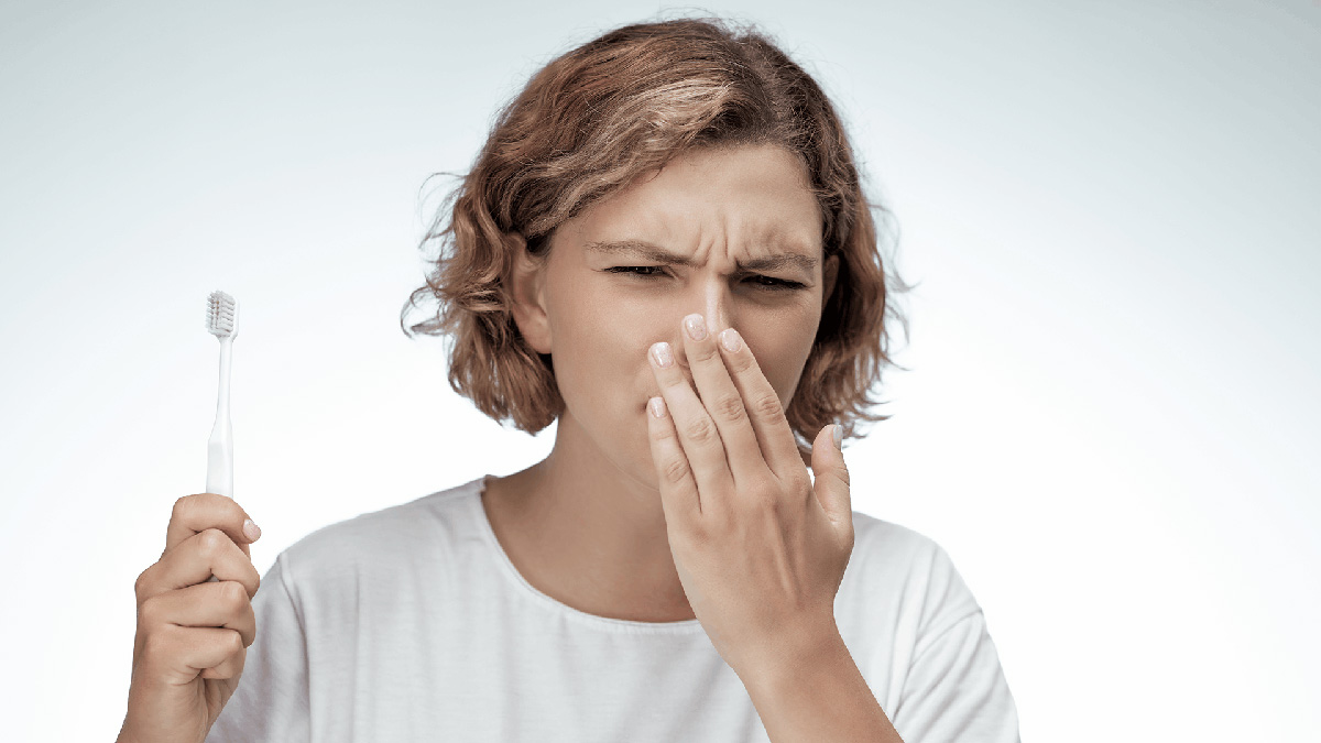 Image of a woman with short hair holding a toothbrush and who has her other hand covering her nose 