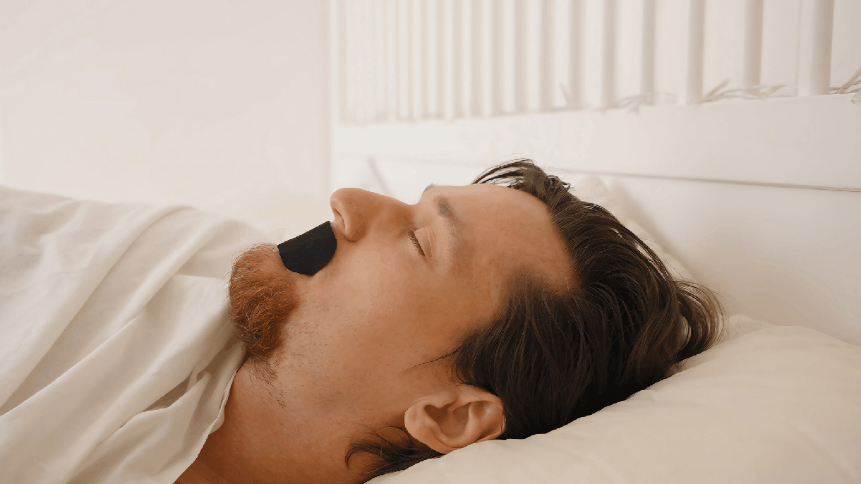 Picture of a man with short brown hair sleeping. He has a black piece of mouth tape over his mouth