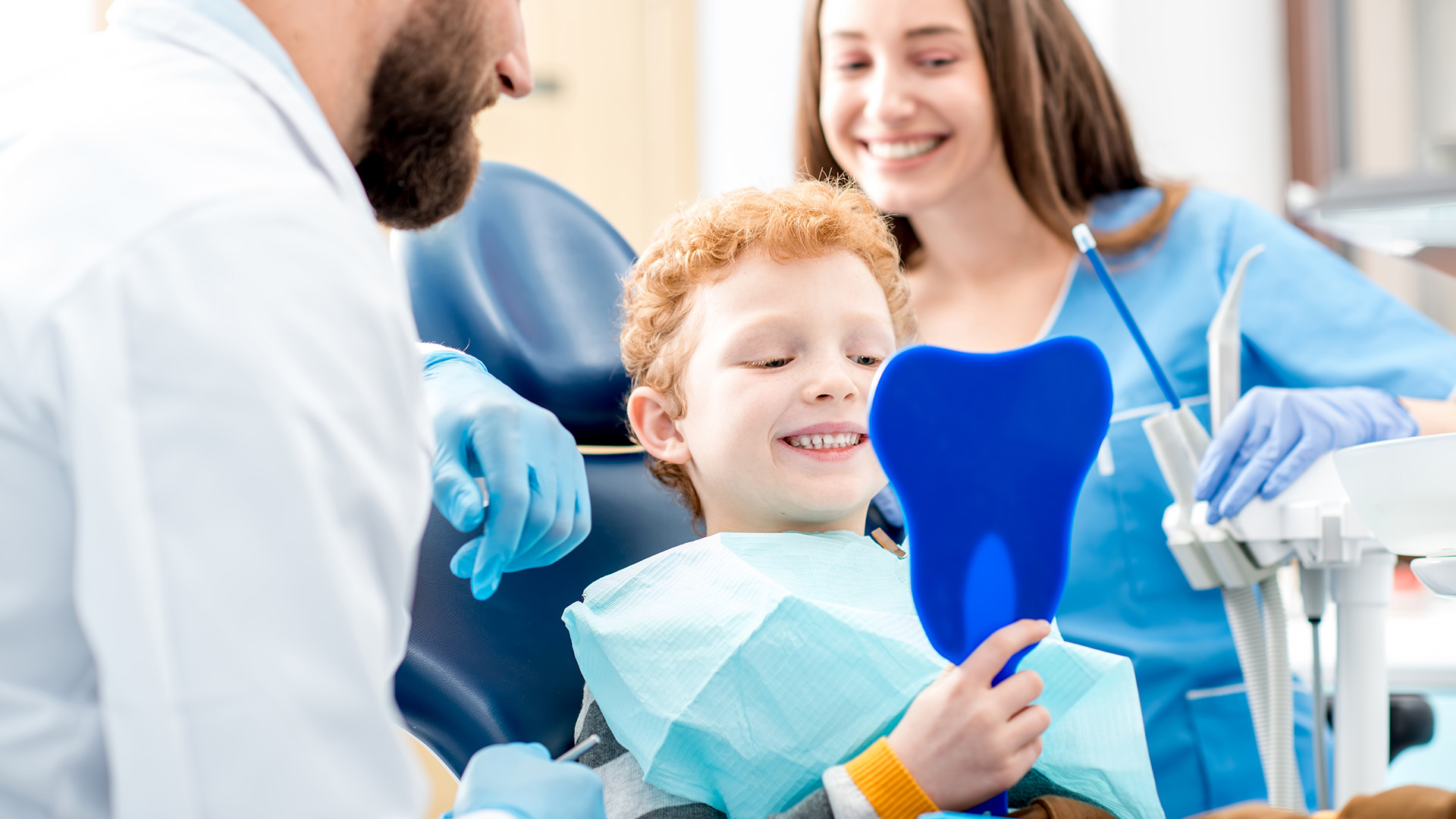 Photo of a young boy with red hair sitting in a dentist chair and holding a blue mirror, smiling while he looks into the mirror